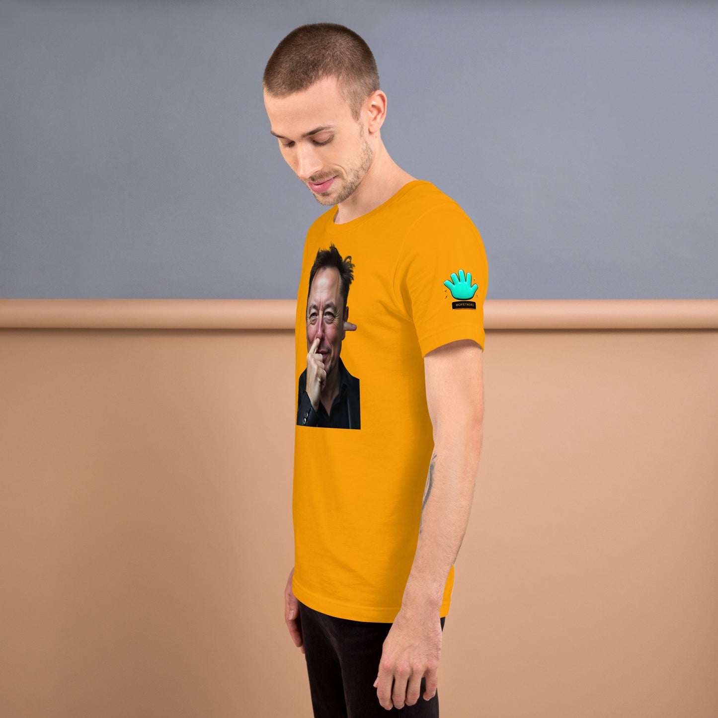 Modelo tatuado mostrando vista lateral de camiseta amarilla Bofetadas 'Celebrities' con logo en manga y impresión frontal con Elon Musk hurgándose la nariz.