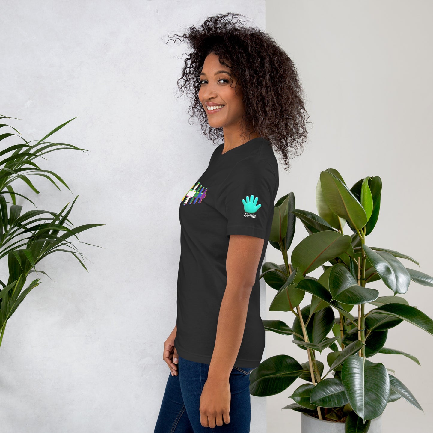 Modelo sonriente con jeans y un cabello voluminoso luce una camiseta gris oscuro de la colección Causas Sociales. Vista lateral, con la impresión en la manga izquierda del logo de la mano azul de Bofetadas.