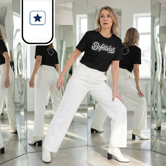 La mujer adopta una postura dinámica y abierta, con las piernas separadas y sin llevar puesta la chaqueta del traje. Viste la camiseta negra con el texto frontal «Bofetadas» y pantalón blanco con botines de tacón medio. A través de los espejos del fondo se aprecia la parte trasera de la prenda, que incluye una ilustración gráfica blanca en la espalda.