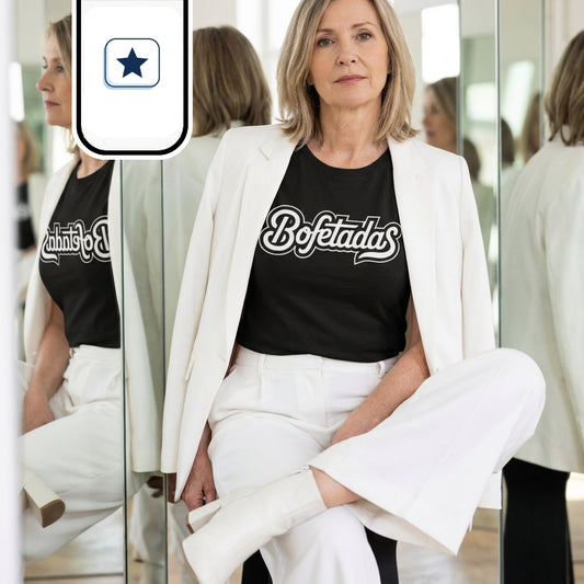 Una mujer madura posa sentada con la pierna cruzada rodeada de espejos. Viste un traje blanco con chaqueta sobre los hombros, botines a juego y una camiseta negra con la palabra «Bofetadas» en el pecho. Su mirada se dirige al frente con una expresión sobria y elegante mientras los cristales reflejan su figura desde distintos ángulos.