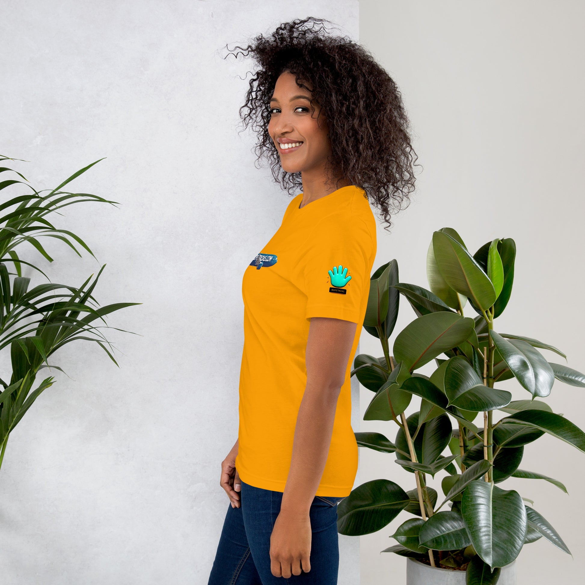 Modelo sonriente con pelo suelto y jeans mostrando la vista lateral de camiseta amarilla de manga corta, marca Bofetadas, colección 'Marca', con el logotipo de una mano azul en la manga.