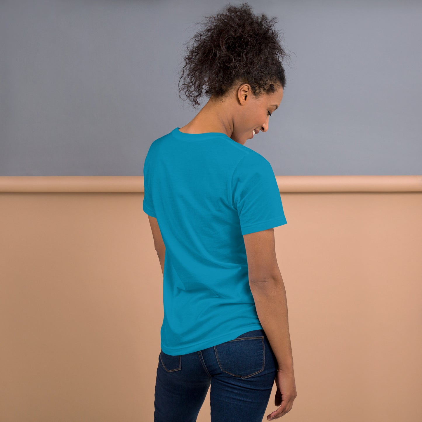 Una modelo sonriente con pelo recogido y jeans mostrando la vista trasera de camiseta azul de manga corta, marca Bofetadas, colección 'Emojis', sin impresión.
