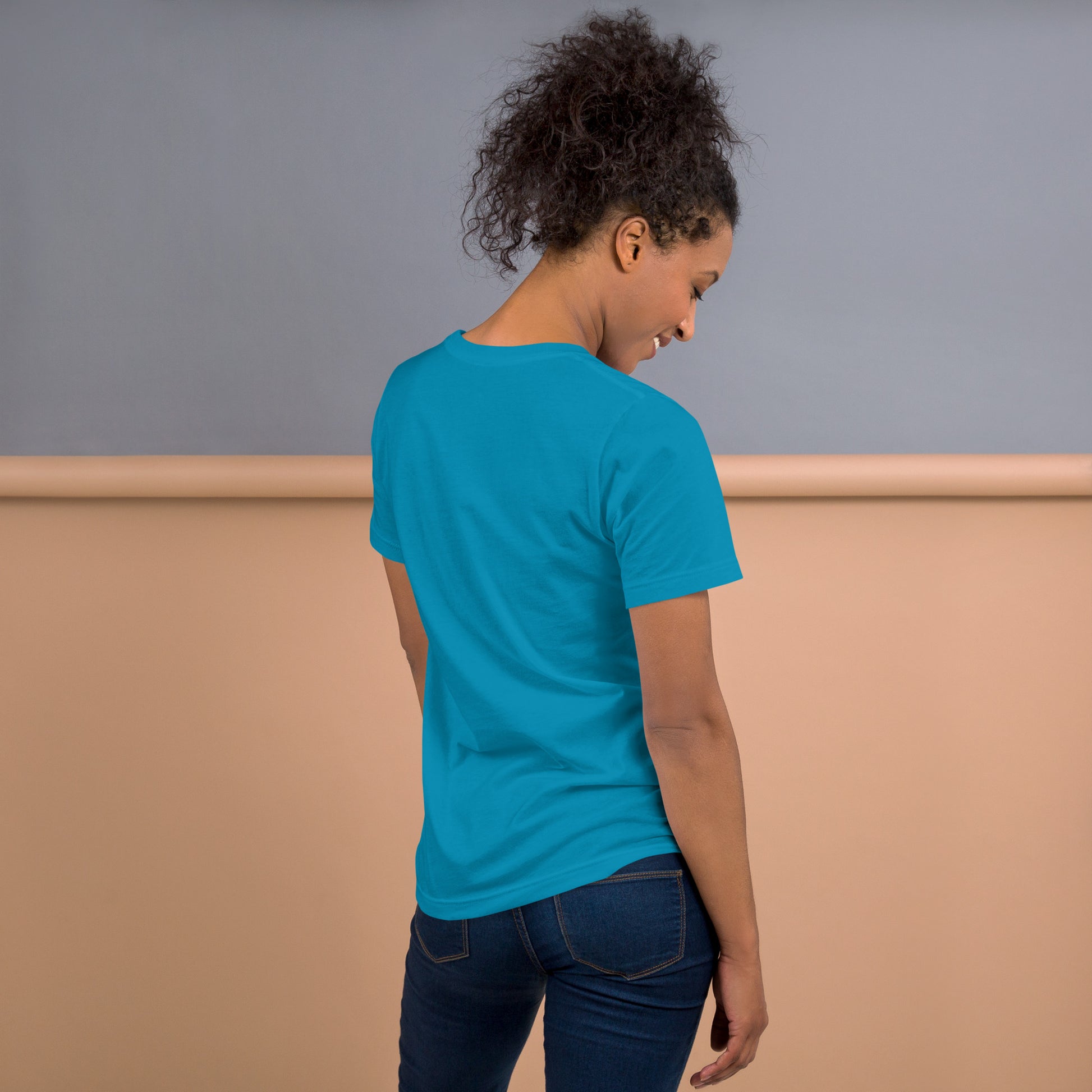 Una modelo sonriente con pelo recogido y jeans mostrando la vista trasera de camiseta azul de manga corta, marca Bofetadas, colección 'Emojis', sin impresión.