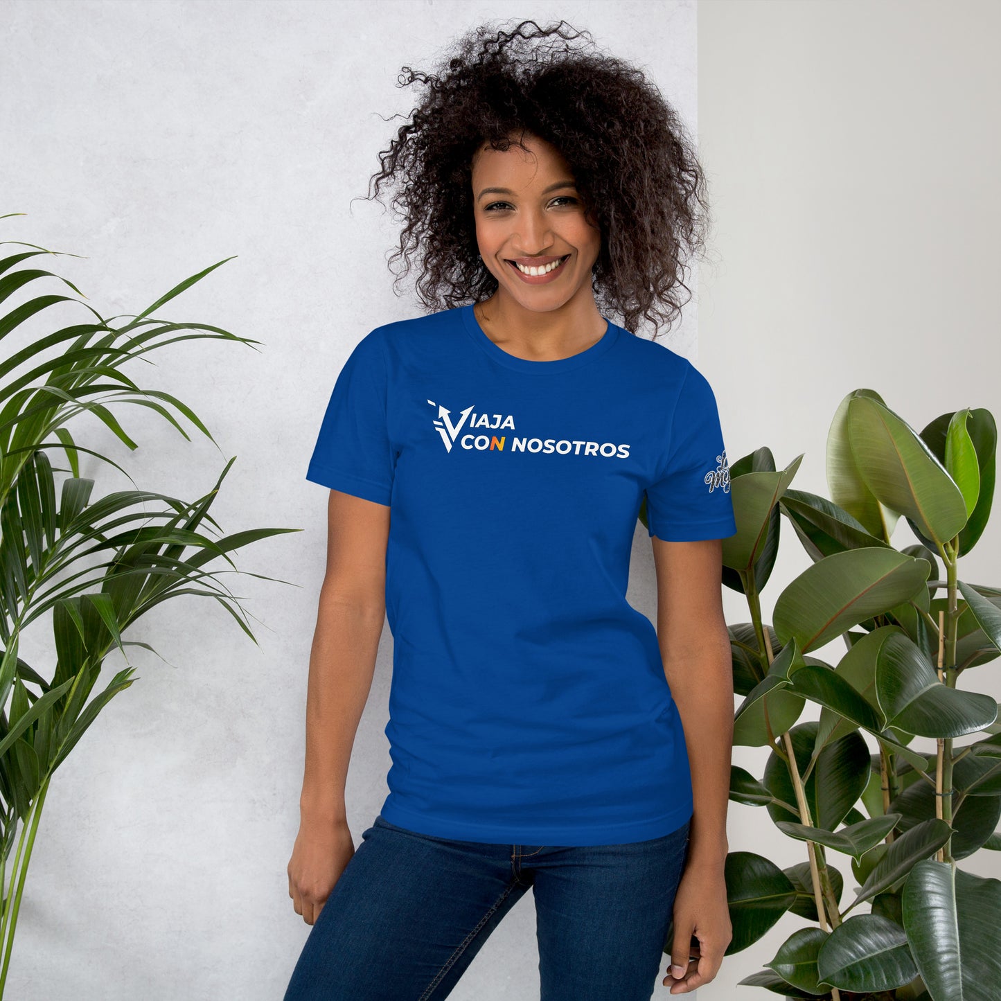 Una modelo sonriente con pelo suelto y jeans luciendo camiseta amarilla de manga corta marca Bofetadas, colección 'Marca', vista frontal. La frase "Viaja con nosotros" con diseño de logotipo moderno en blanco. La letra "V" se estiliza como dos flechas audaces que sugiere movimiento continuo y la letra "N" de con es de color naranja.