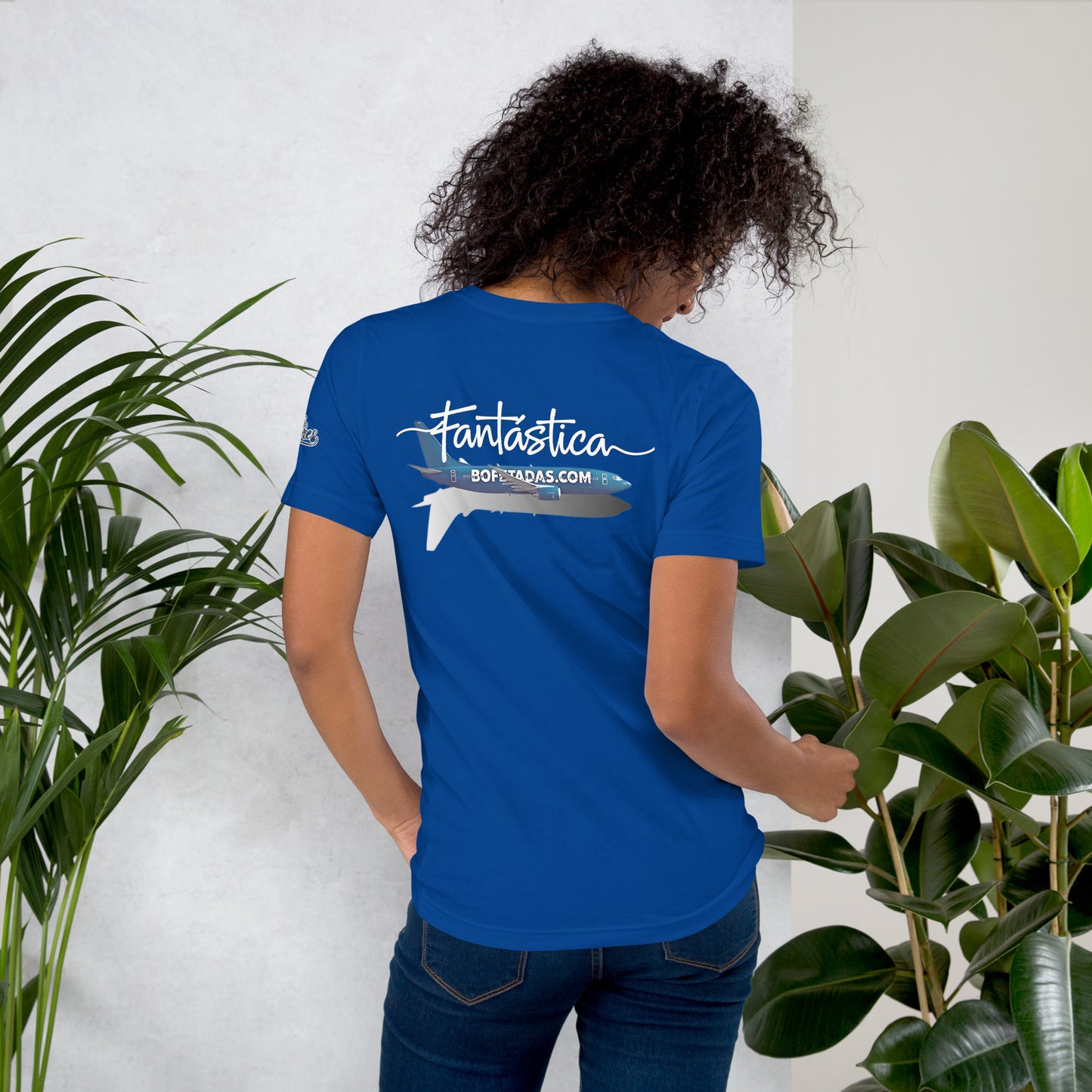 Modelo con pelo suelto y jeans mostrando la vista trasera de camiseta amarilla de manga corta, marca Bofetadas, colección 'Marca', con  la impresión de un avión azul y su sombra  con texto impreso en el fuselaje 'bofetadas.com'. También la palabra "Fantástica" de un solo trazo en blanco encima del avión.