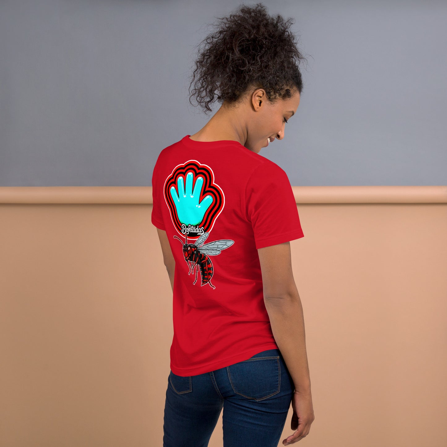 Modelo sonriente con jeans y el pelo recogido luce camiseta roja colección 'marca', vista trasera, con la impresión de el logo de la mano azul con la marca envueltos por halos rojos y negros acompañados de una avispa roja.