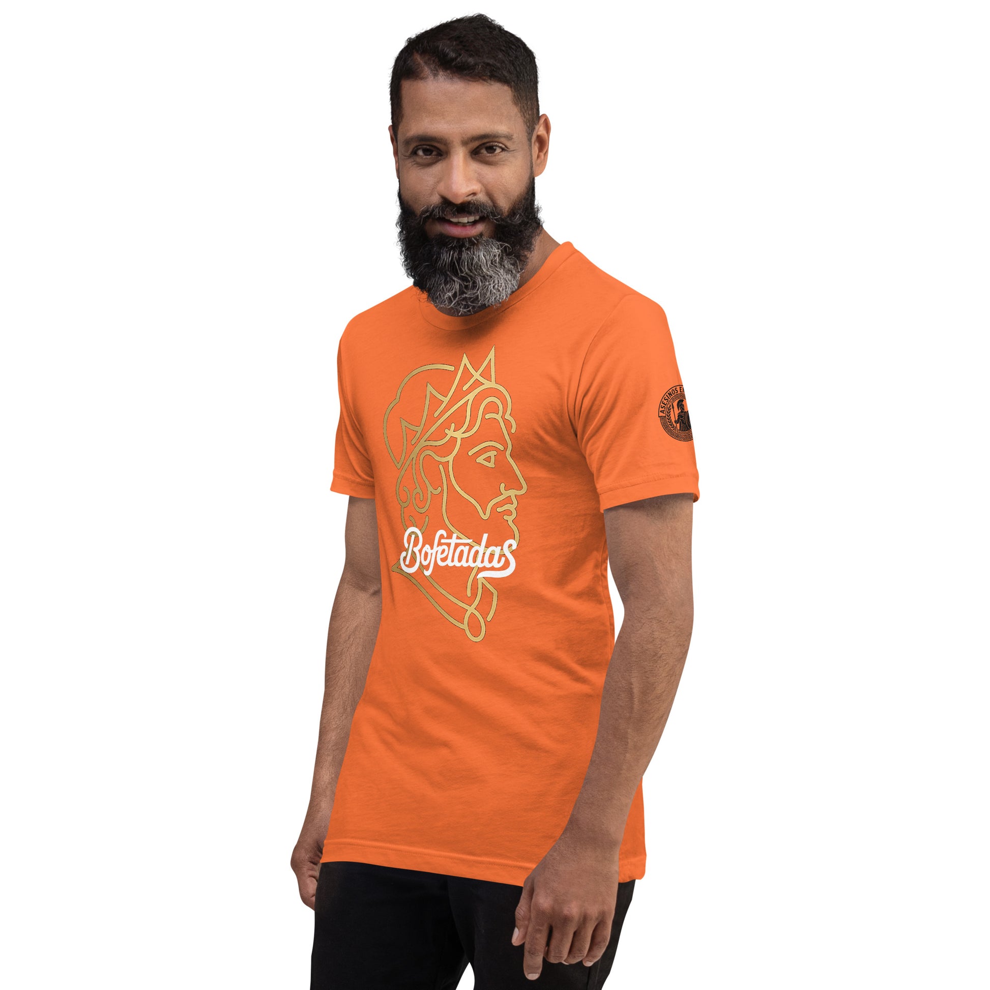 Un modelo con barba y pelo corto luce una camiseta naranja de Bofetadas de la colección Asesinos en serio. Tiene impreso en el frente una ilustración lineal de el rostro de Alejandro Magno y la tipografía de Bofetadas. Se aprecia en la manga izquierda el logotipo de la nueva colección.