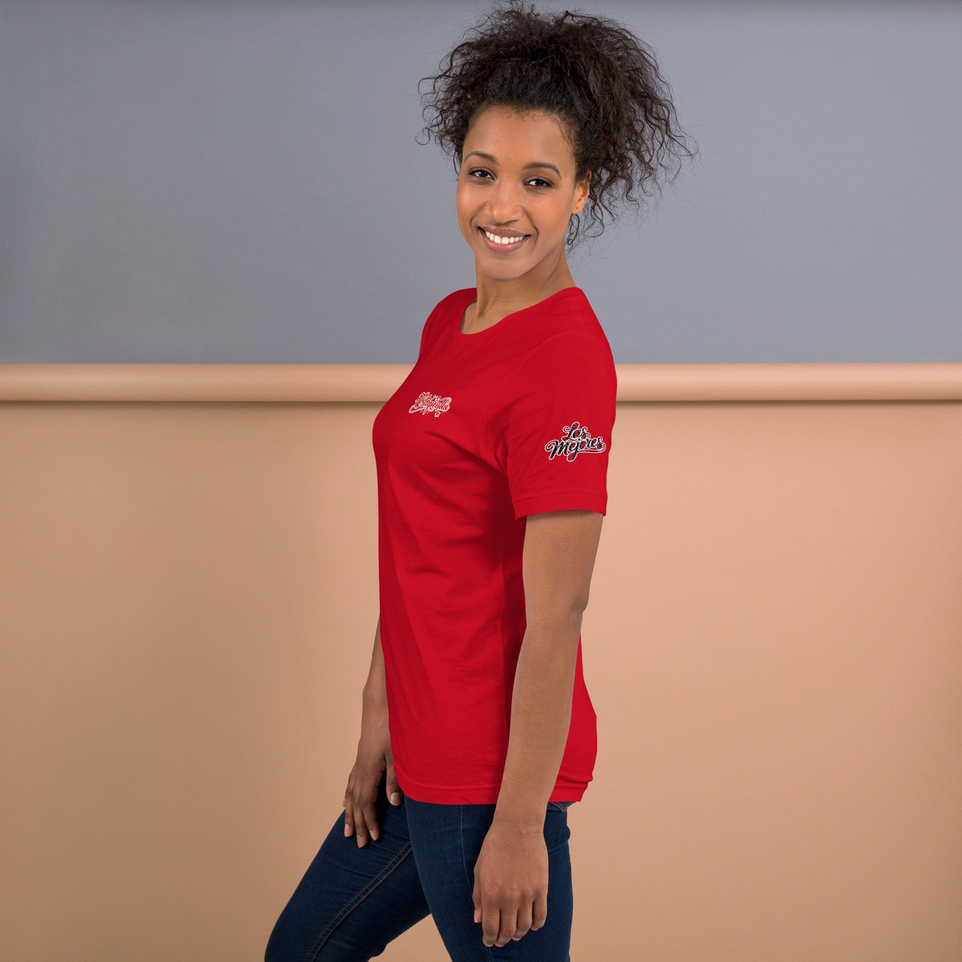 Modelo sonriente con jeans luce camiseta roja colección 'marca', vista lateral, con el texto 'Estupenda' en el pecho y 'Las mejores' con estilo caligráfico inspirado en Pokras Lampas en la manga izquierda.
