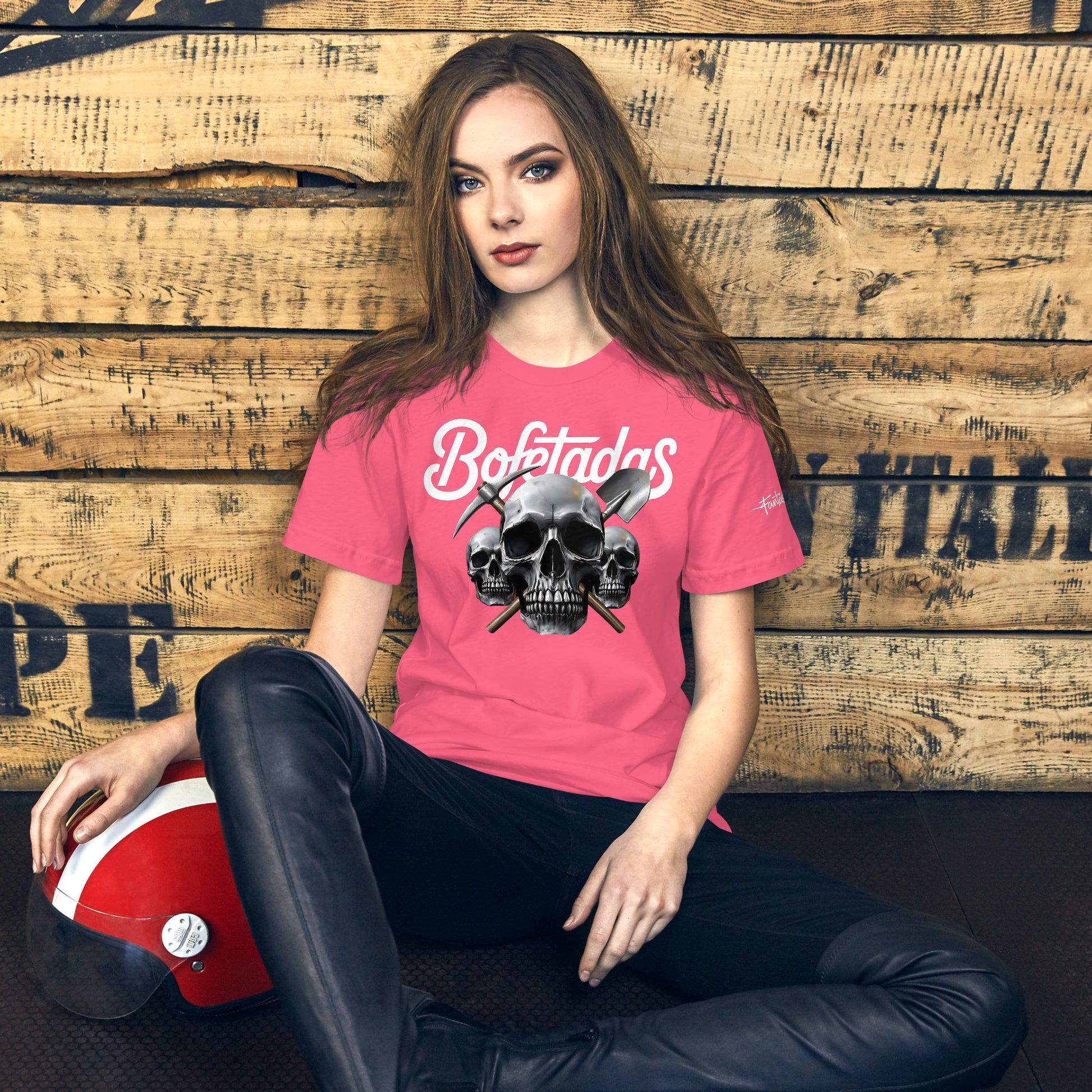 Una modelo sentada en su habitación luce una camiseta rosa de manga corta marca Bofetadas, colección 'Calaveras', con la impresión de tres calaveras, una pala y un pico cruzados completando la bandera pirata. Y el texto "Bofetadas".